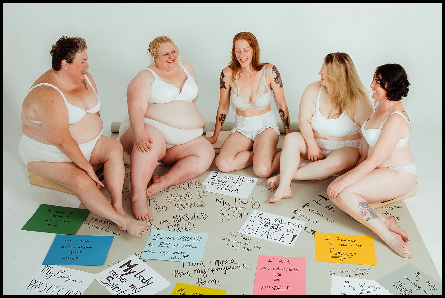 Five women of diverse body types and sizes sit together in white underwear, smiling and laughing. This Nashville boudoir empowerment shoot surrounds them with handwritten affirmations on colorful paper, celebrating body positivity and self-love.