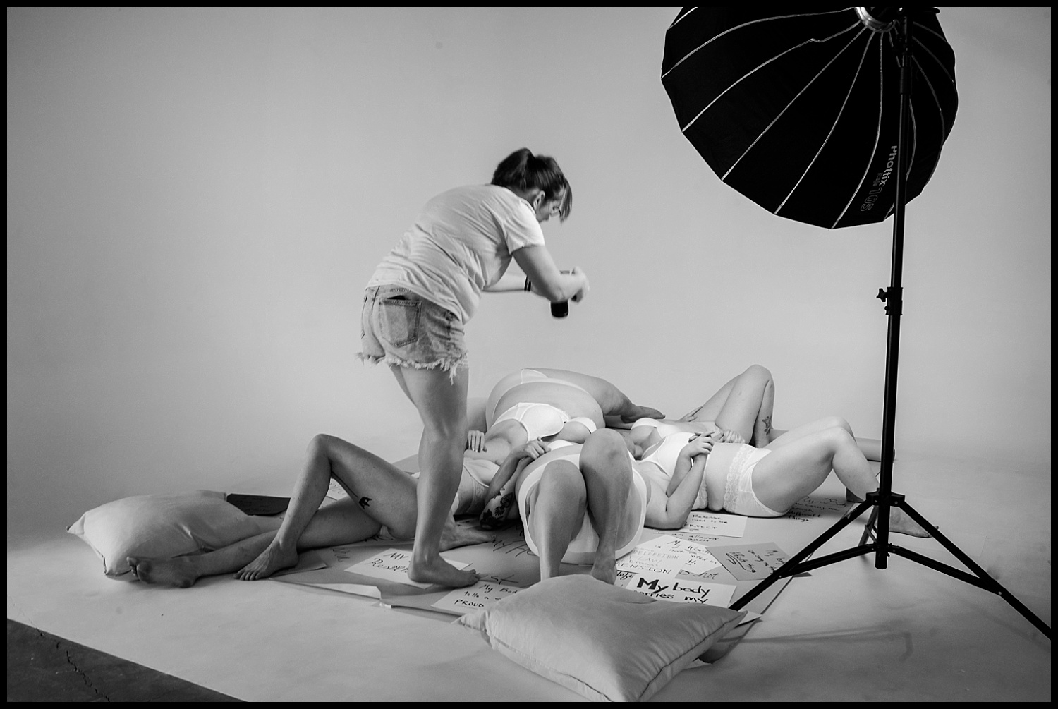 A photographer leans over three people lying on the floor, surrounded by pillows and handwritten signs, capturing a Nashville boudoir empowerment shoot in a studio with a large softbox light overhead. The photo is black and white.