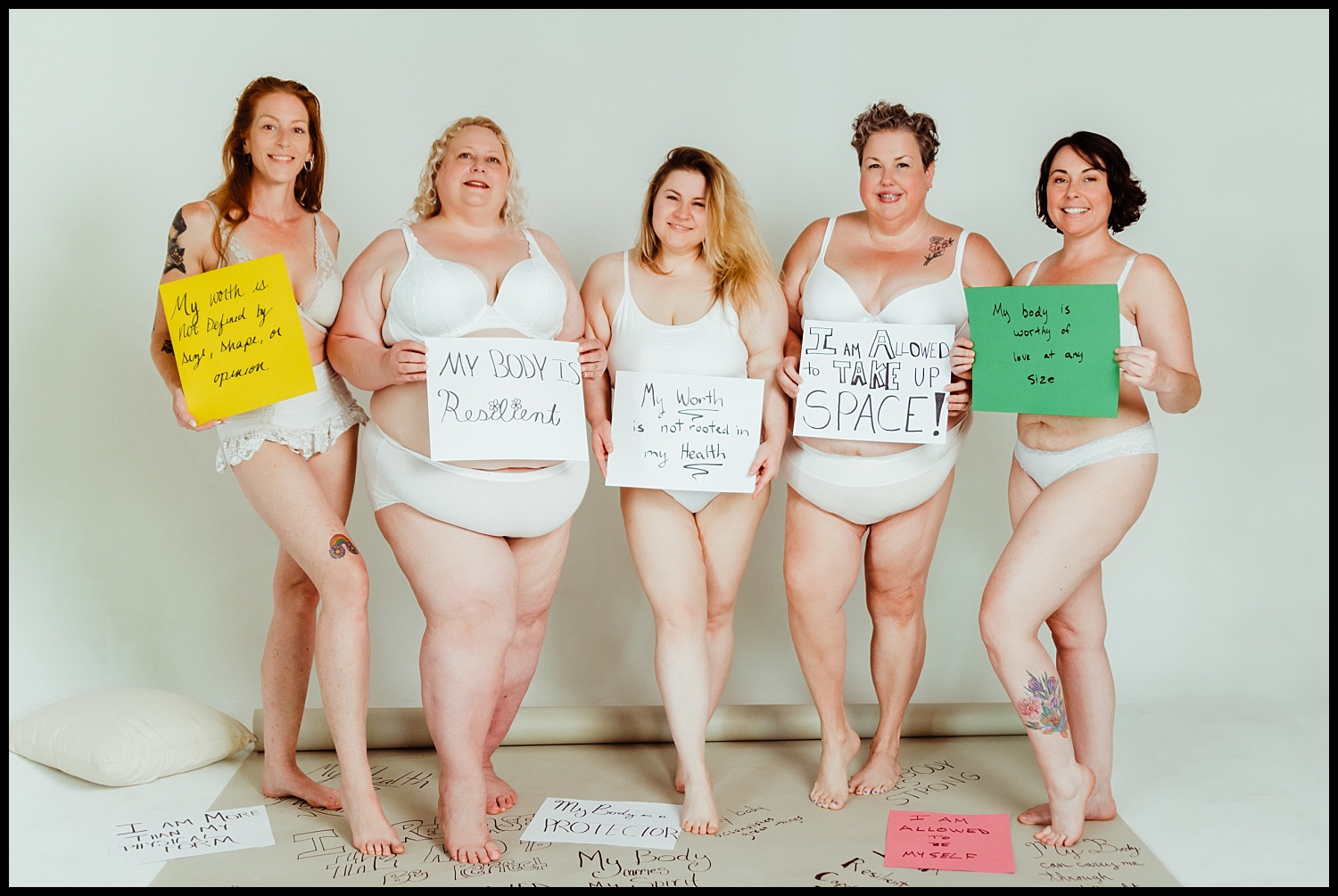Five women of diverse body types, wearing white underwear, stand together holding colorful handwritten signs promoting self-love and empowerment. They are smiling and standing on a white backdrop for a Nashville Boudoir Empowerment Shoot.