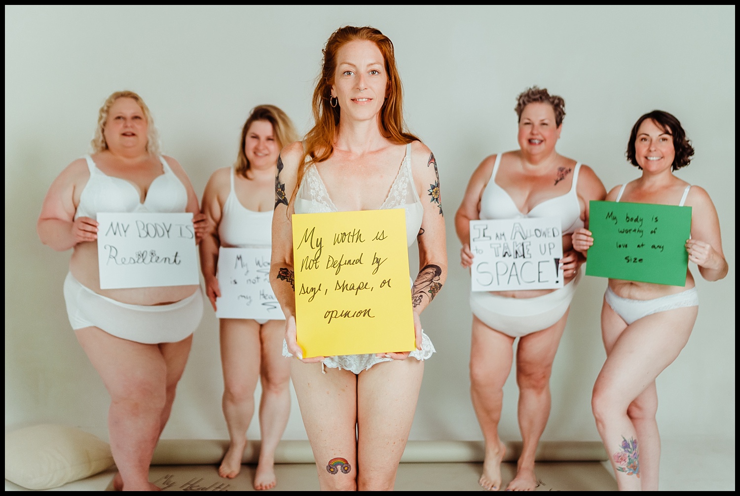 Five women in white underwear stand confidently, each holding handwritten signs with body positivity messages, promoting self-love and self-worth, against a plain background—a powerful Nashville boudoir empowerment shoot.