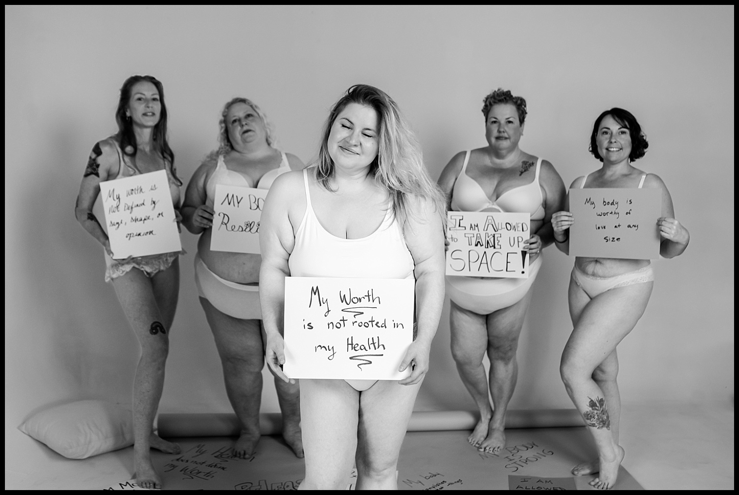 Five women of diverse body types stand together in underwear, each holding a sign promoting body acceptance and self-worth. They smile confidently in a bright, minimal studio setting. The Nashville boudoir image is black and white.