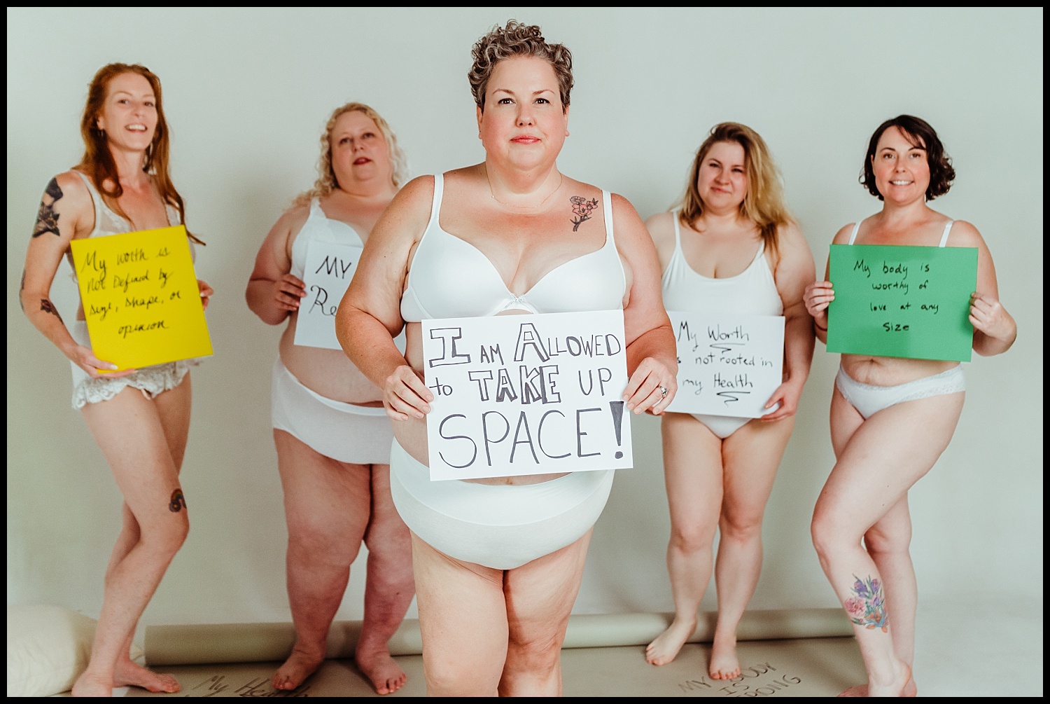 Five women of different body types stand confidently in white underwear, holding handwritten signs promoting self-love and body positivity against a plain light background—a beautiful moment of body acceptance captured by a Nashville boudoir session.