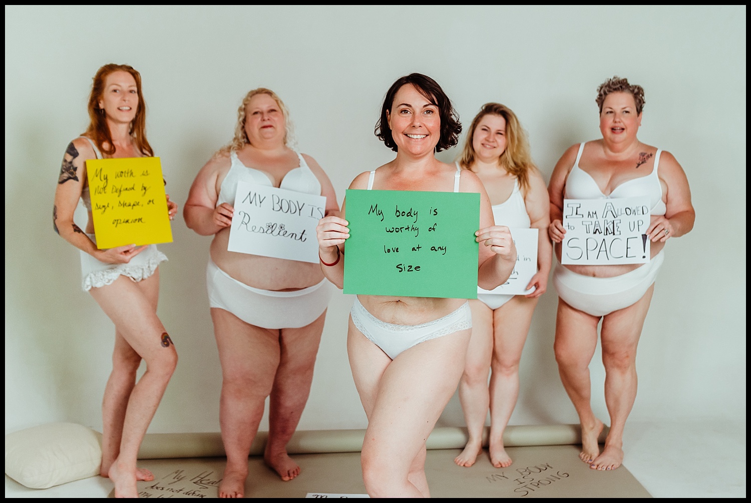 Five women of diverse body types, wearing white underwear, stand confidently holding signs with empowering messages about self-love and body positivity against a plain background. They smile and embrace self-acceptance and boudoir empowerment.