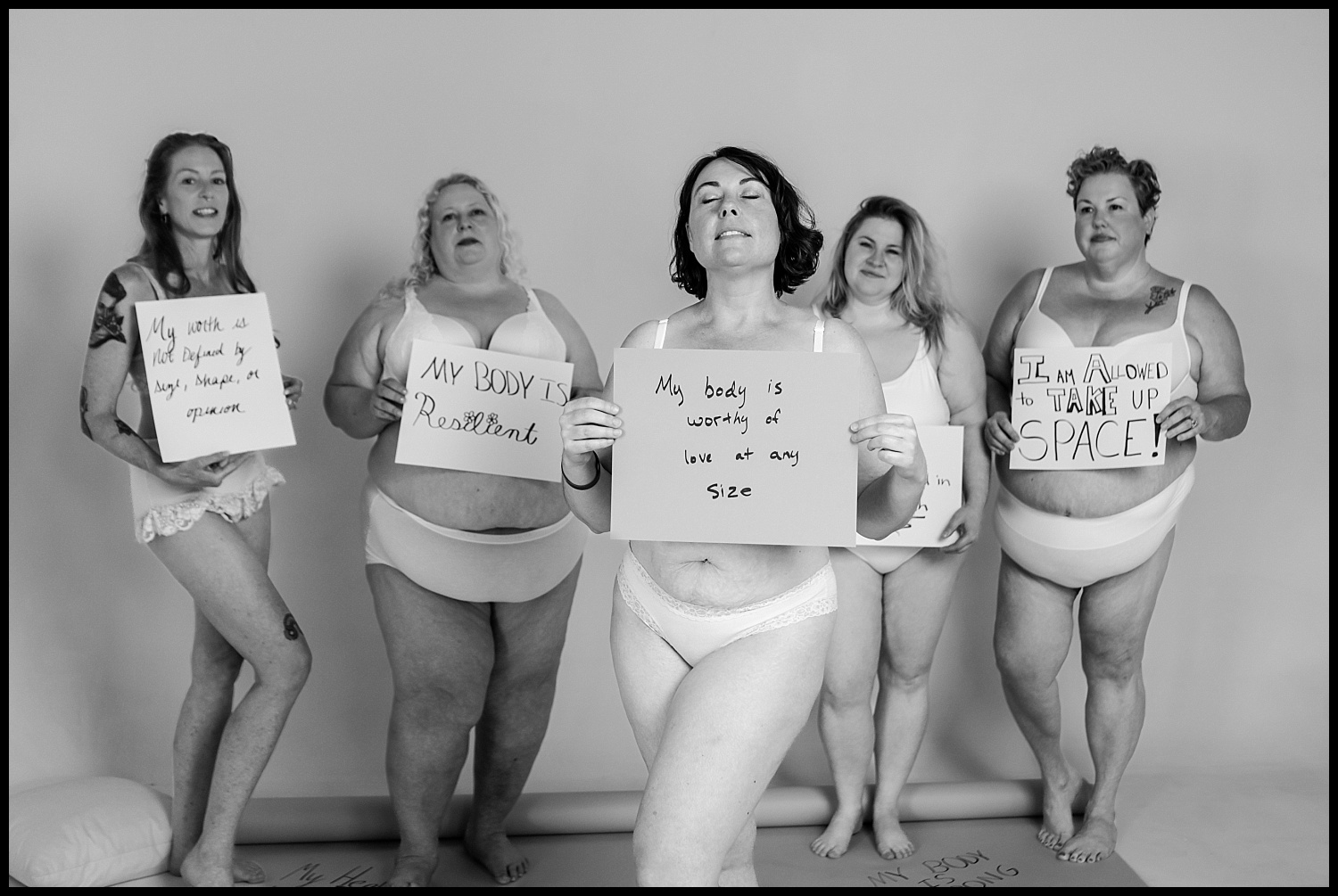 Five women in white underwear stand confidently, each holding a handwritten sign with body-positive messages of self-love and body acceptance, against a plain background. The black and white photo captures the empowering spirit of a Nashville Boudoir session.