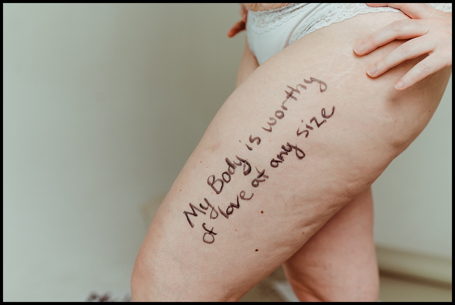 A close-up of a person’s upper thigh with stretch marks, wearing white underwear. “My body is worthy of love at any size” is written on their skin in marker, celebrating self-love and body acceptance.
