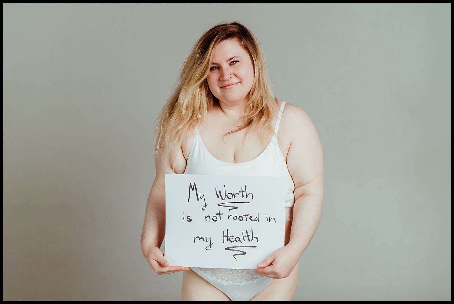 A woman in white underwear holds a sign that says, My worth is not rooted in my health. She stands against a plain, light-colored background, radiating confidence and embodying self-love and body acceptance.