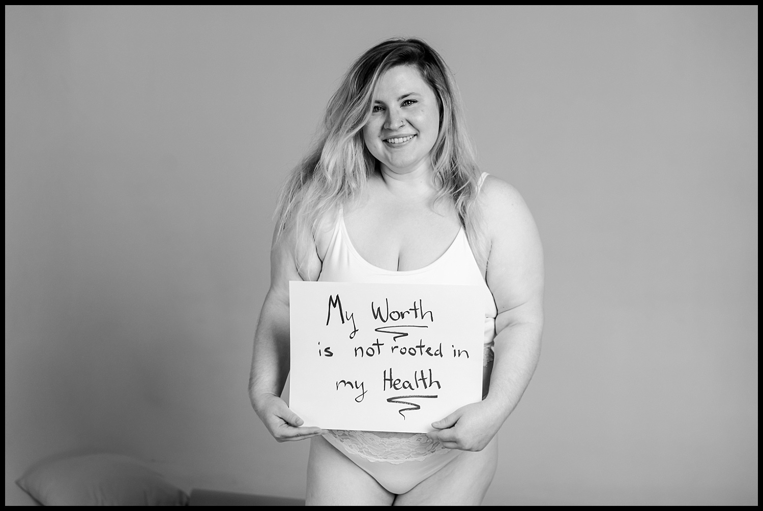 A woman wearing a white tank top and underwear smiles while holding a sign that reads, My worth is not rooted in my health. This Nashville boudoir scene embraces self-love and body acceptance against a plain, neutral background.