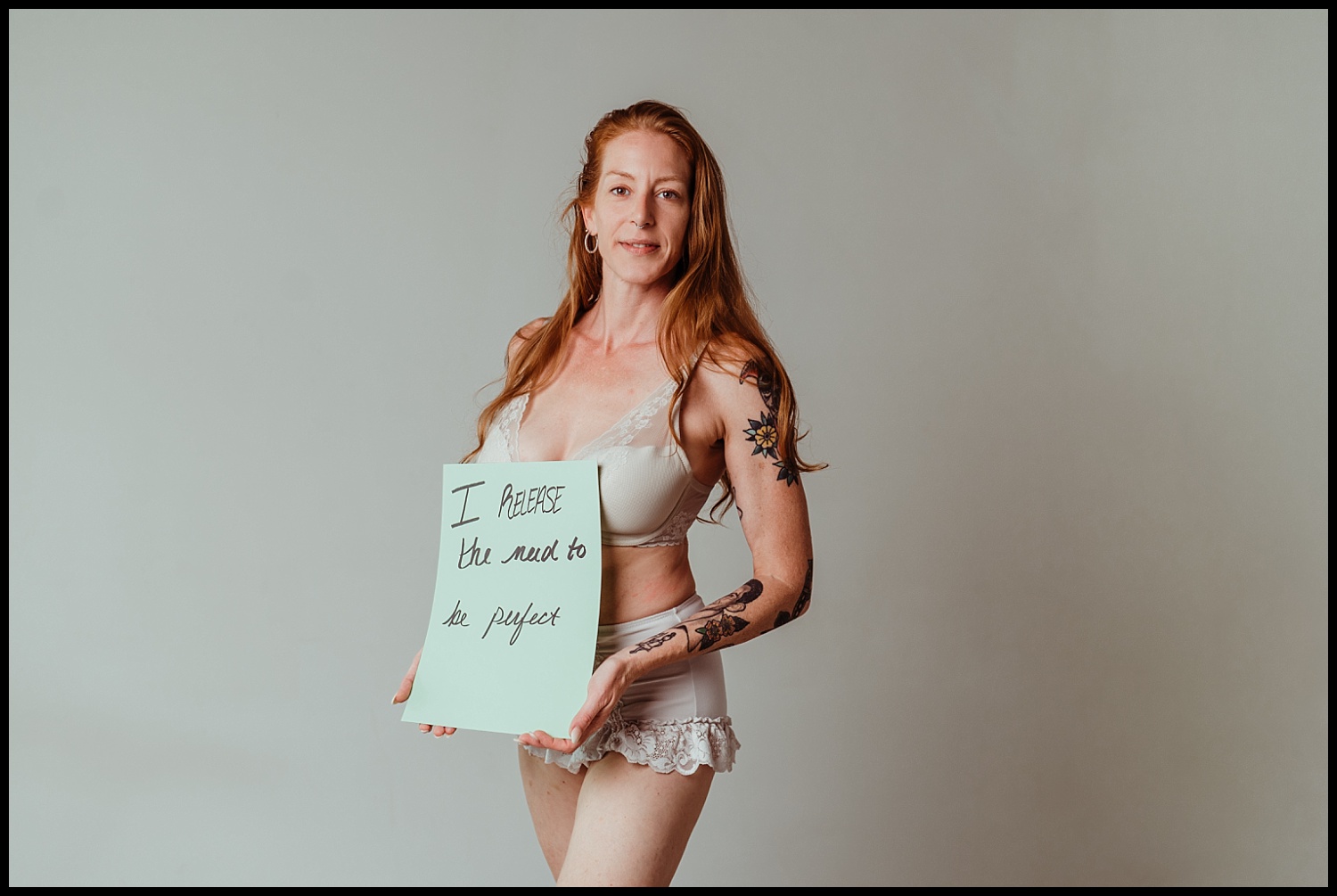 A woman with long red hair and tattoos stands confidently in light-colored lingerie against a plain background, holding a sign that reads, “I release the need to be perfect.” This Nashville boudoir empowerment shoot radiates self-love.