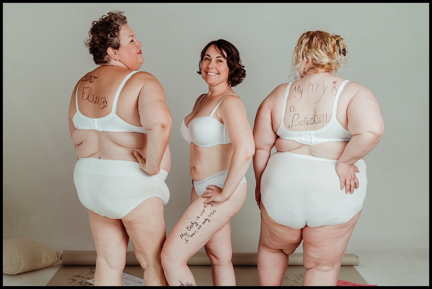 Three women in white underwear stand together, showing body acceptance messages written on their skin. They face the camera and away, smiling confidently in a bright, neutral setting—a true Nashville boudoir vibe of self-love and empowerment.