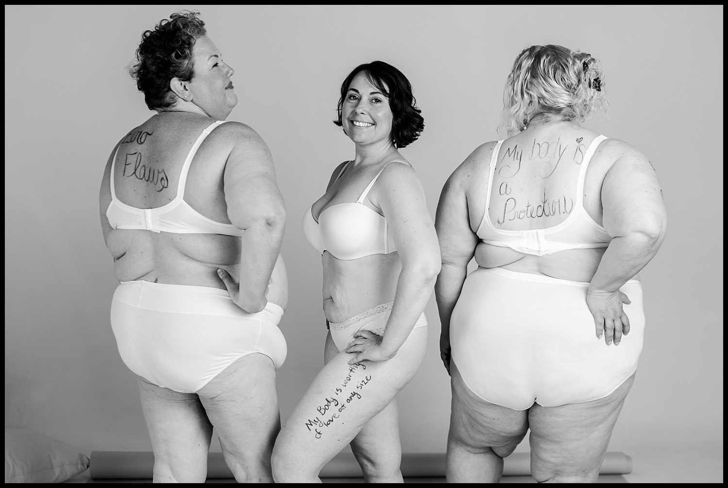 Three women wearing white bras and underwear pose confidently with positive messages on their skin. This black and white Nashville boudoir image celebrates self-love and body acceptance, as all three women smile, facing different directions.