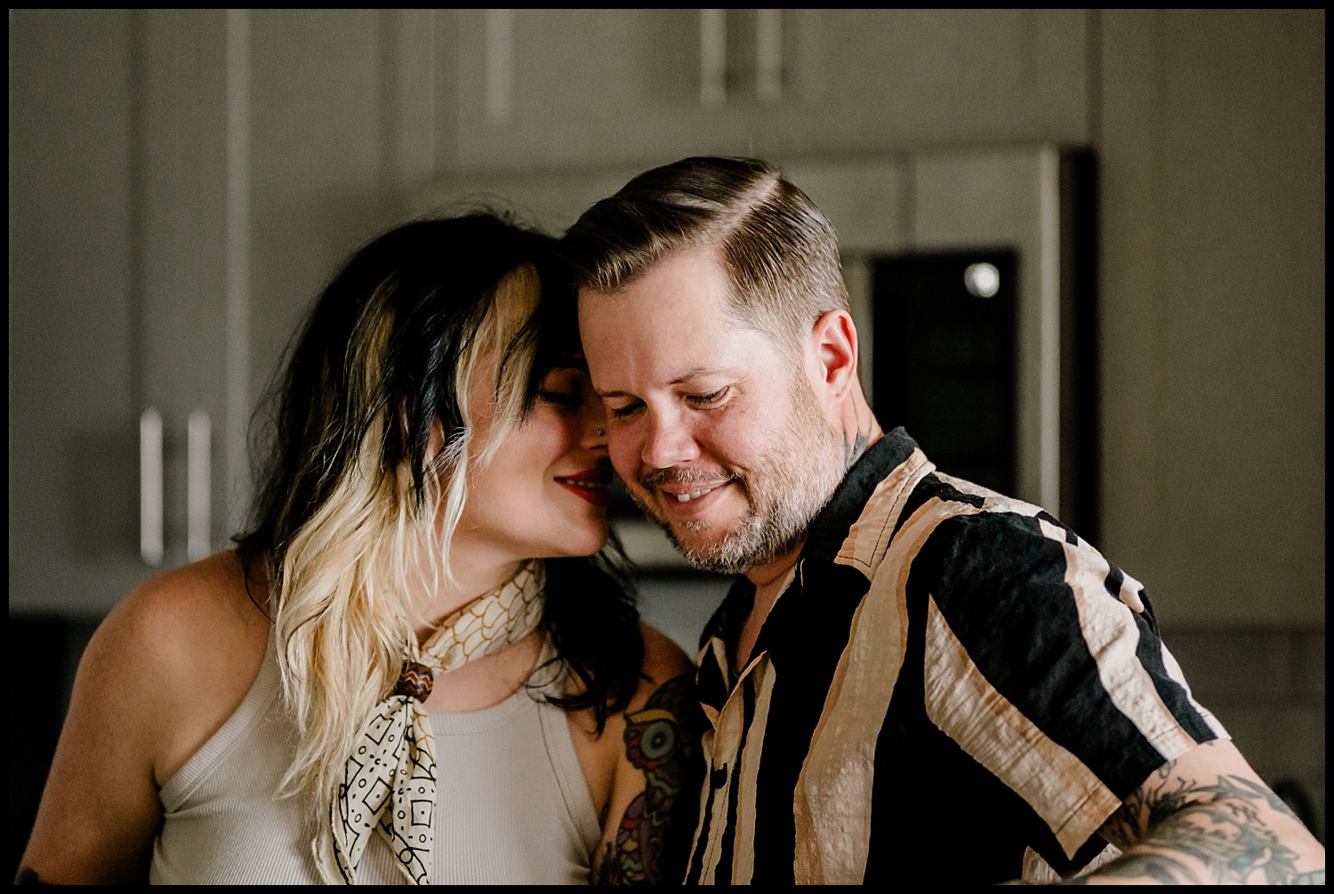 A smiling couple stands close together in their Nashville kitchen during an in-home couples session. The woman leans in, touching her face to the mans, both looking happy and relaxed amid cabinets and a microwave.
