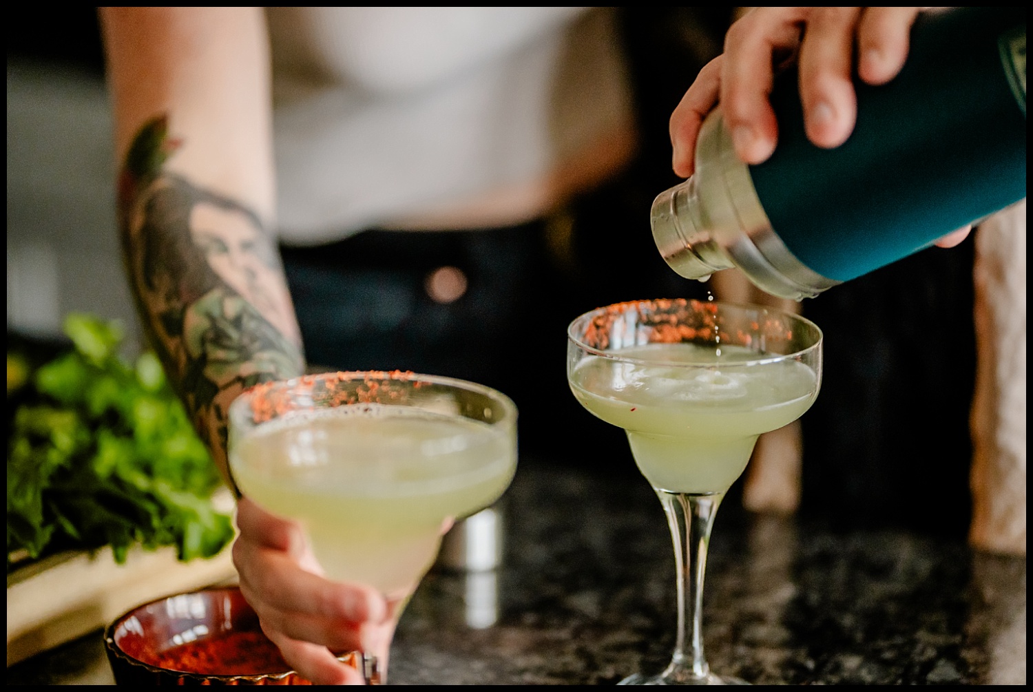 A person with a tattooed arm pours a light green cocktail from a shaker into a chili powder–rimmed glass; another similar drink sits nearby on a dark countertop, perfect for enjoying before tacos during a Nashville couples session. Fresh herbs are visible in the background.