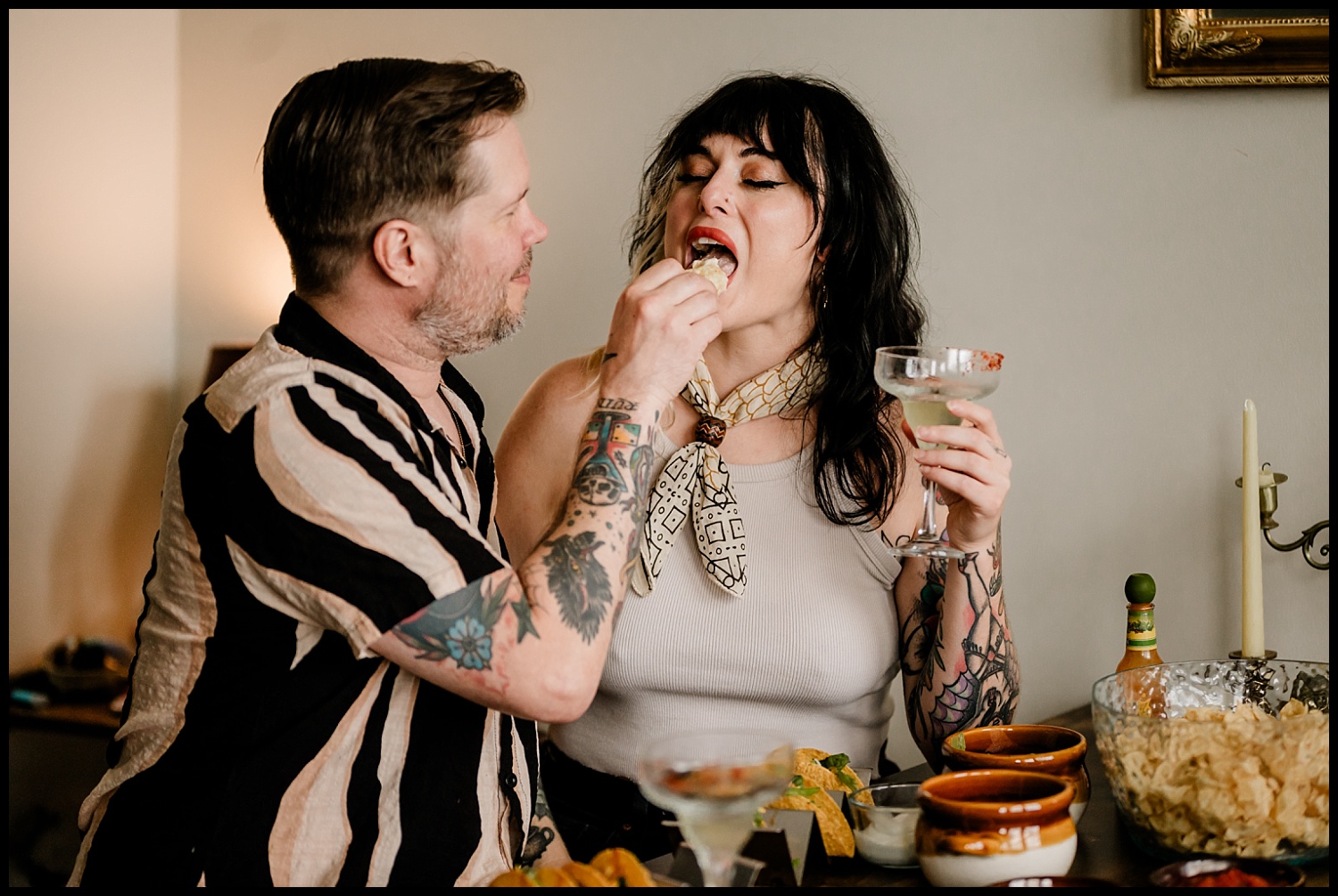 A tattooed man feeds a tortilla chip to a tattooed woman holding a cocktail, both smiling at a lively table with chips, salsa, and drinks. This Nashville couples session captures their fun, casual vibe.