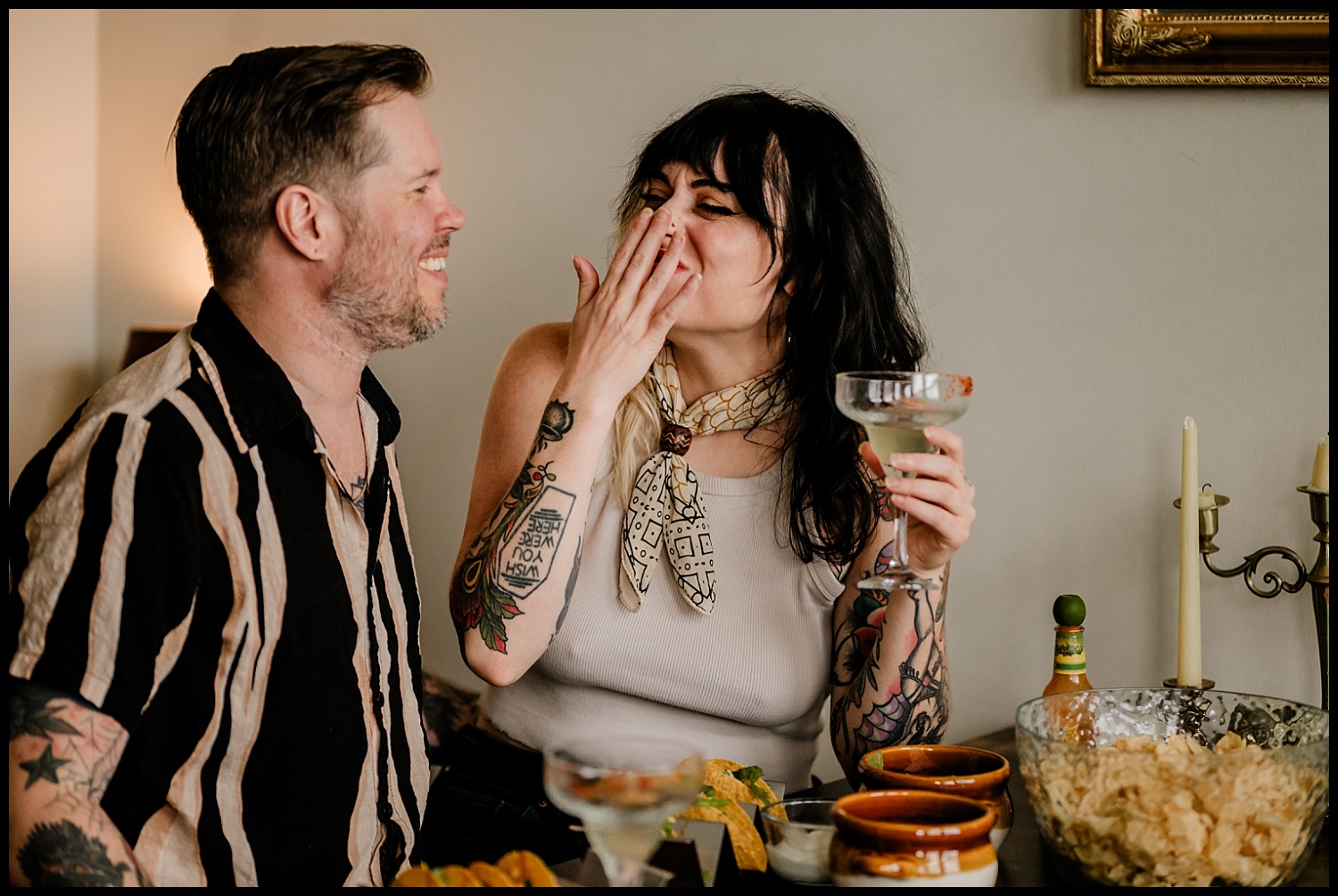 A smiling couple enjoys drinks, tacos, and chips during a Nashville couples session. The woman laughs over her cocktail while the man looks at her lovingly. The atmosphere feels casual and joyful, capturing genuine moments together.