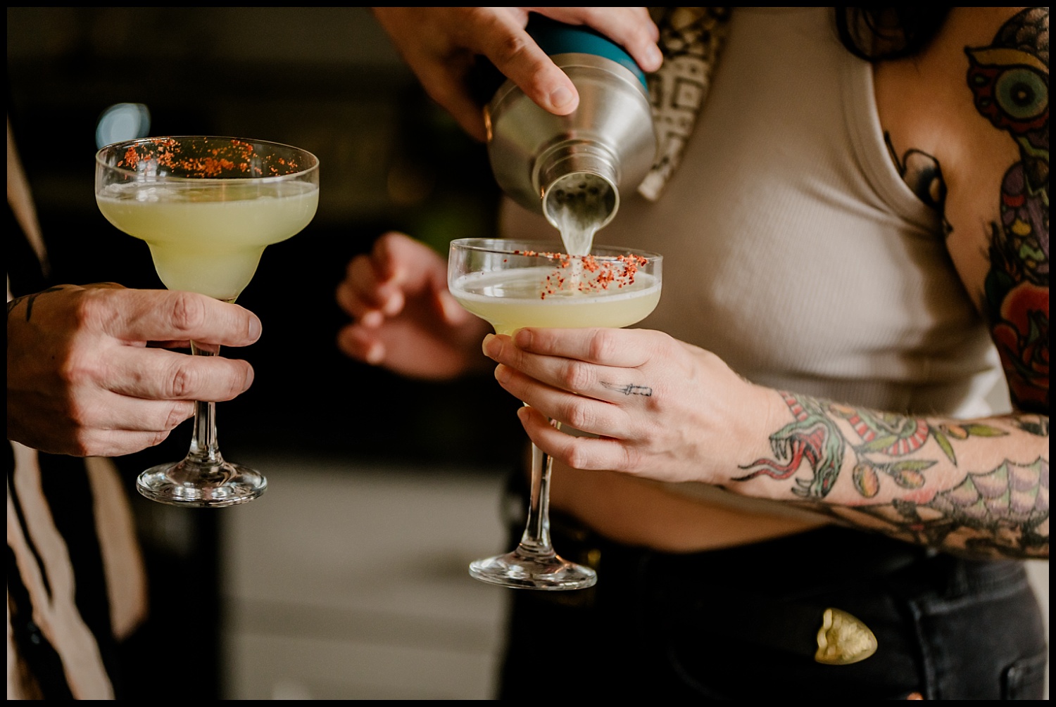 Two people with tattoos toast tequila cocktails with a chili rim during a couples session. One pours from a shaker, both in casual attire. The focus is on their hands and vibrant drinks, perfect for pairing with tacos.
