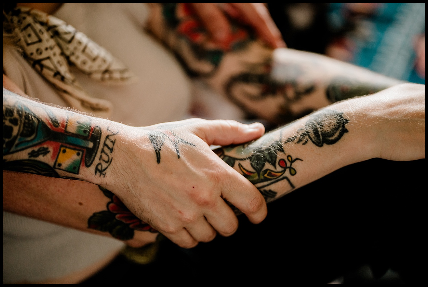 A close-up of two tattooed arms, one person gently holding the others forearm during a couples session. The tattoos feature colorful designs, bold text, and nods to tacos and tequila, with both people sporting fair skin.