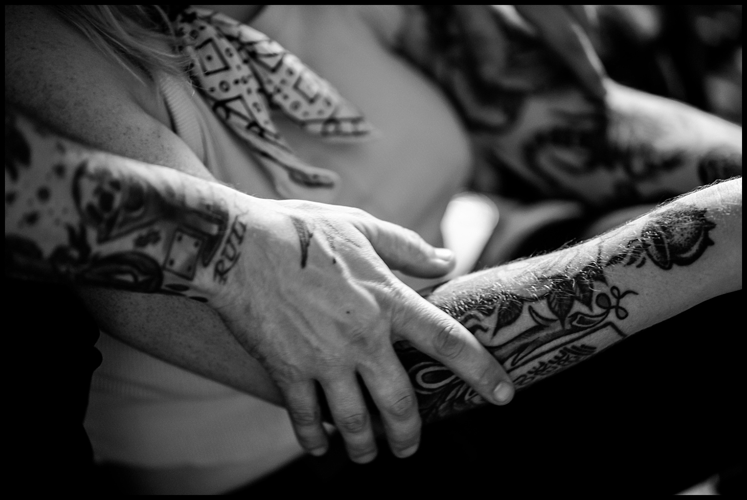 Black and white photo of two tattooed arms intertwined during an in-home couples session, with one hand gently resting on the others forearm. The scene captures an intimate and comforting moment between the individuals in Nashville.