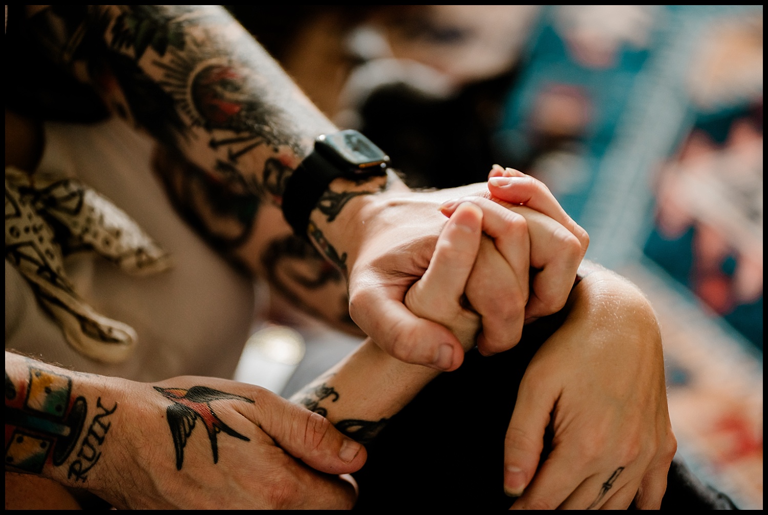 A close-up of two tattooed arms holding hands tightly during an in-home couples session, showing colorful tattoos and a black smartwatch on one wrist, with a blurred background.