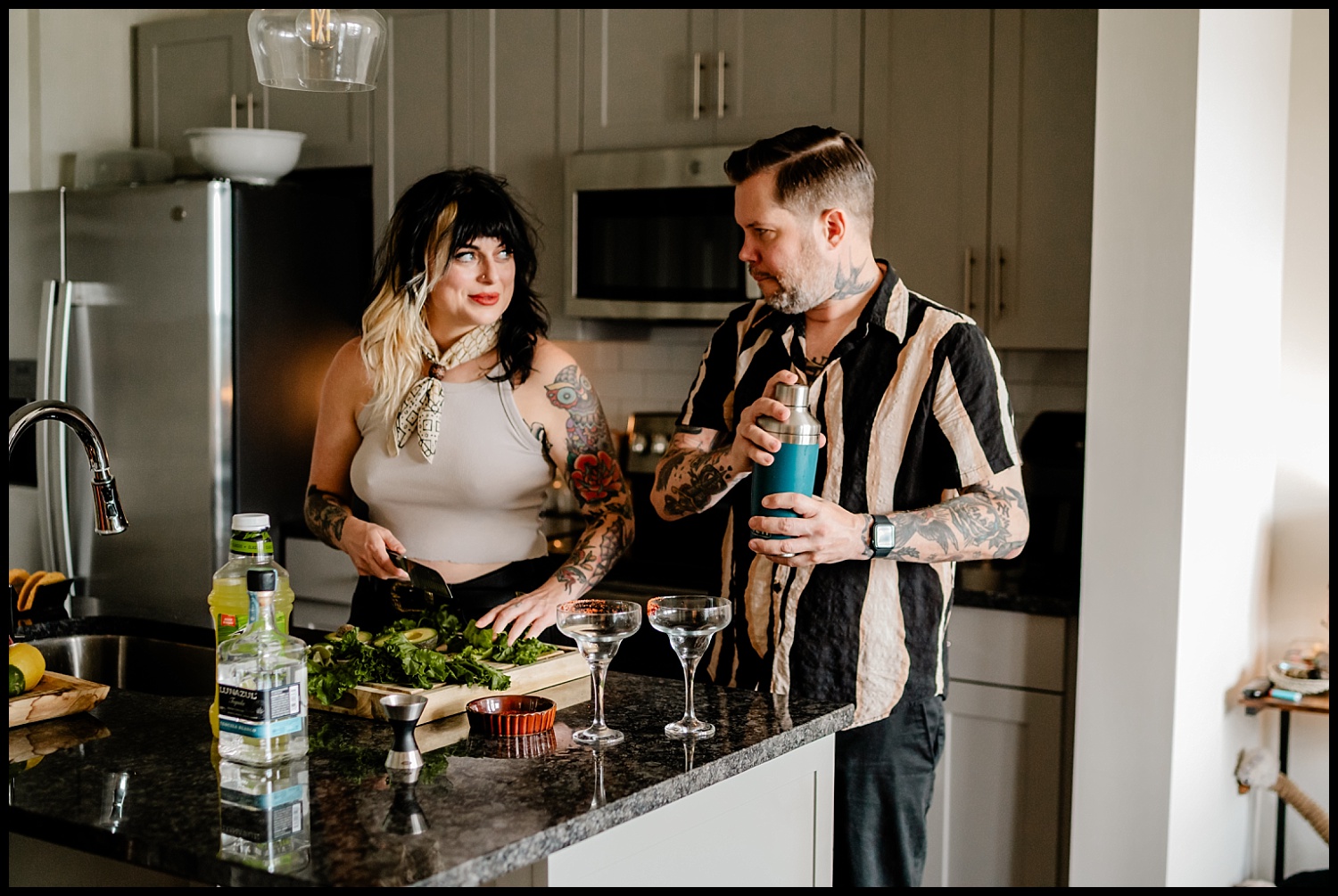 A woman chops greens on a kitchen counter while a man beside her shakes a cocktail mixer. With tattoos and smiles, this Nashville couples session captures their joy in an authentic in-home photography setting.