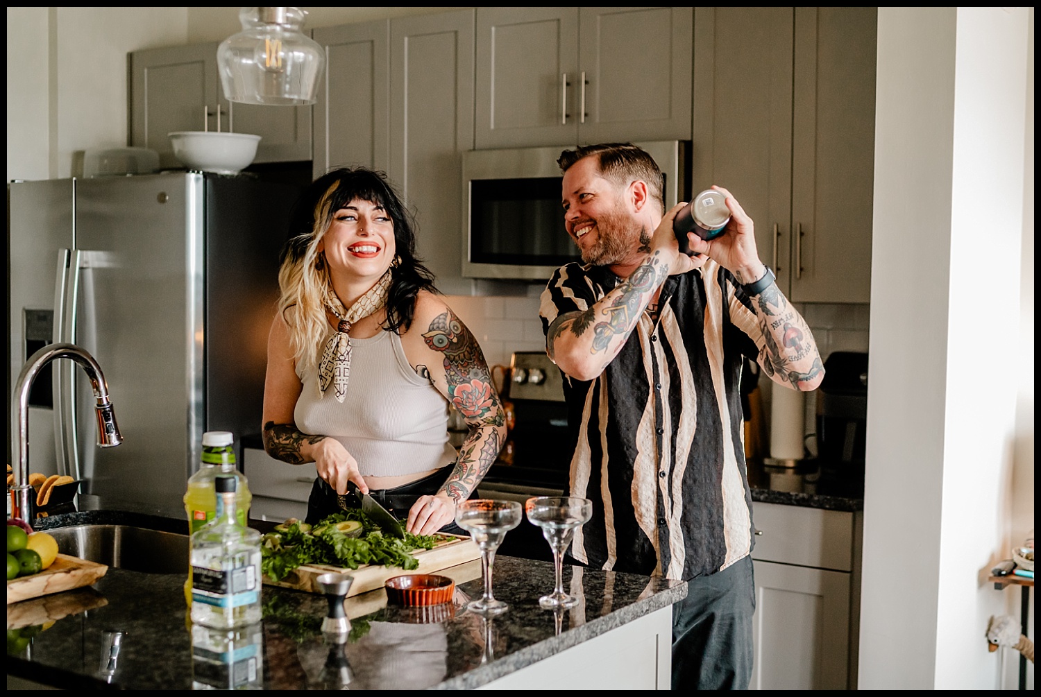 Two people with tattoos smile and prepare drinks in a modern Nashville kitchen during an in-home couples session; one chops herbs on a cutting board while the other shakes a cocktail shaker. Bottles, glasses, and limes are on the countertop.