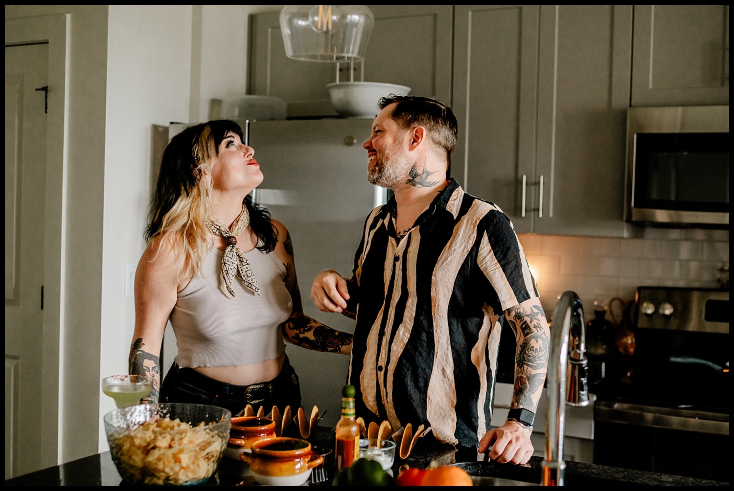 A smiling couple with tattoos stands in a modern kitchen, chatting and laughing. The counter in front of them is set with chips, salsa, drinks, and tacos, suggesting they are preparing for a fun couples session or gathering.