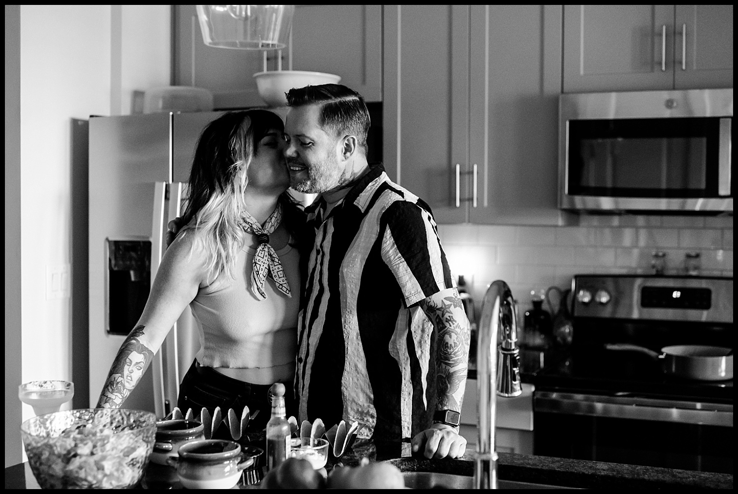 A woman kisses a smiling man on the cheek in a modern kitchen during an intimate couples session. Both have tattoos and casual clothes. Food and utensils are visible on the counter in front of them. The black and white scene captures relaxed in-home photography.