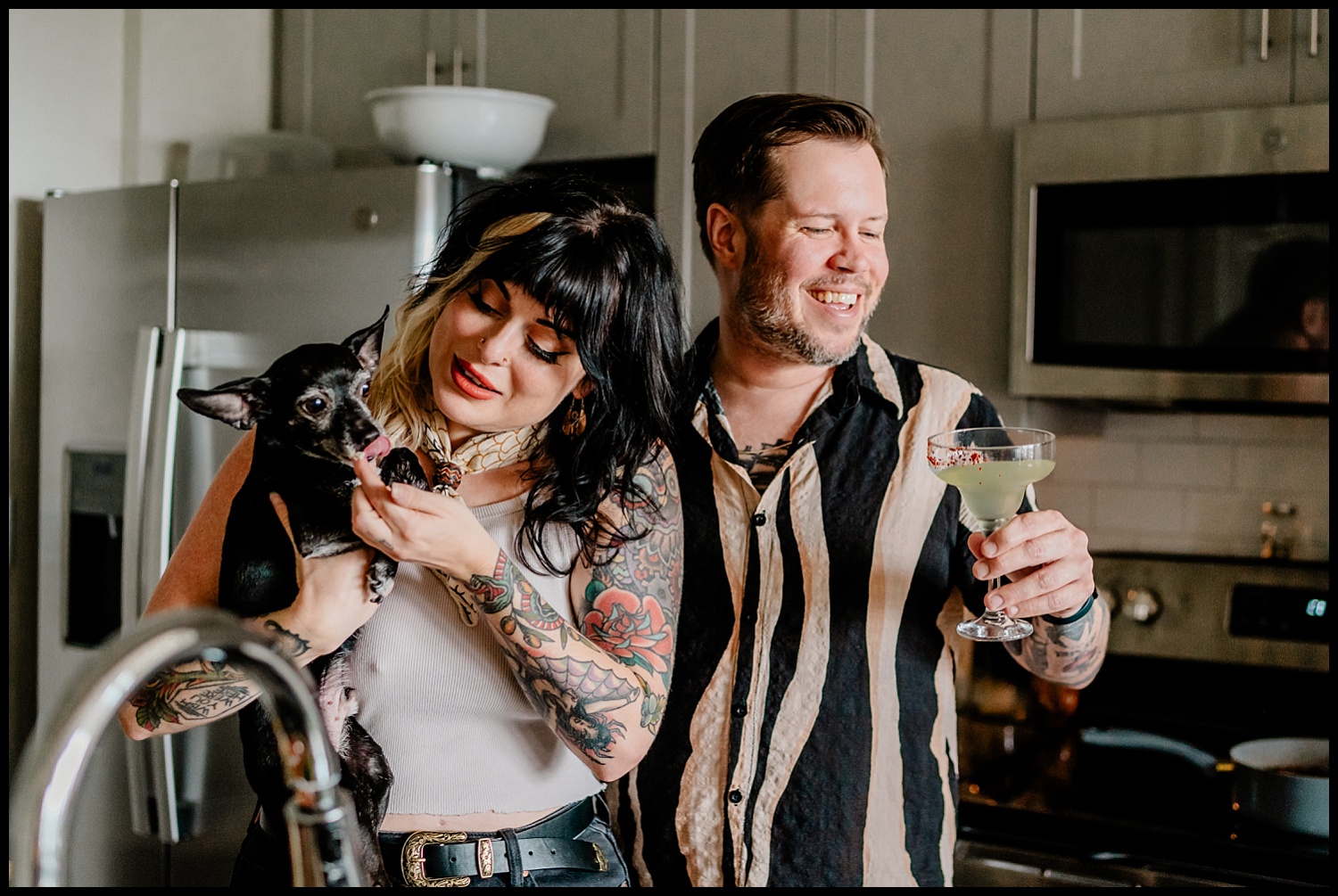 A woman with tattoos holds a small black dog in a kitchen, smiling as the dog licks her face during an in-home couples session. A man beside her, also smiling, holds a cocktail glass. Stainless steel appliances are in the background.