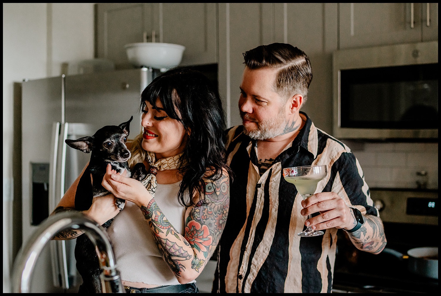 A woman holding a small black dog smiles as a man with a drink stands beside her in their modern Nashville kitchen. Both have tattoos and appear happy and relaxed during this cozy in-home couples session.