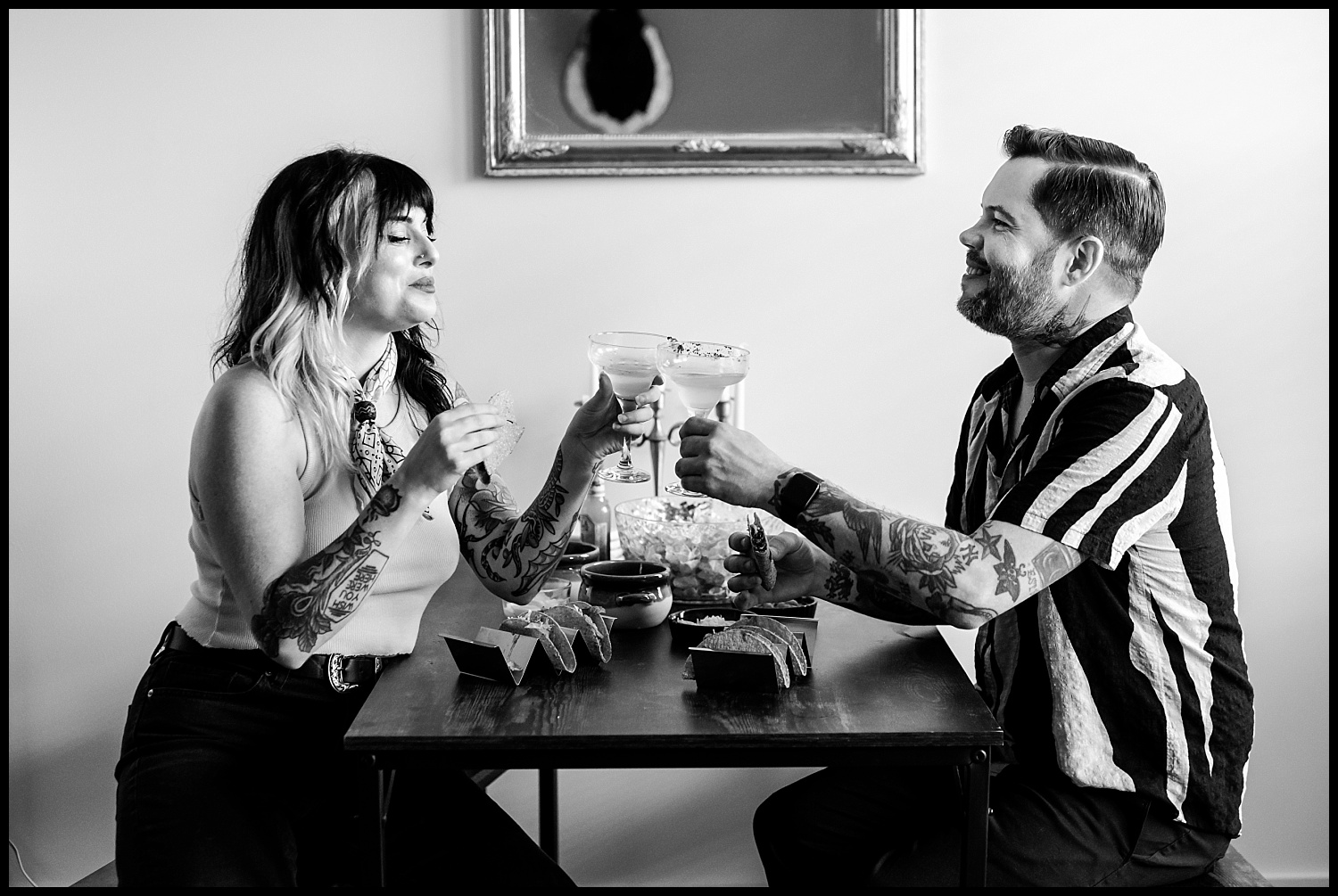 A woman and a man with tattoos sit at a small table during an in-home couples session, smiling and clinking cocktail glasses over tacos and bowls of food. A framed mirror hangs on the wall behind them. The image is in black and white.