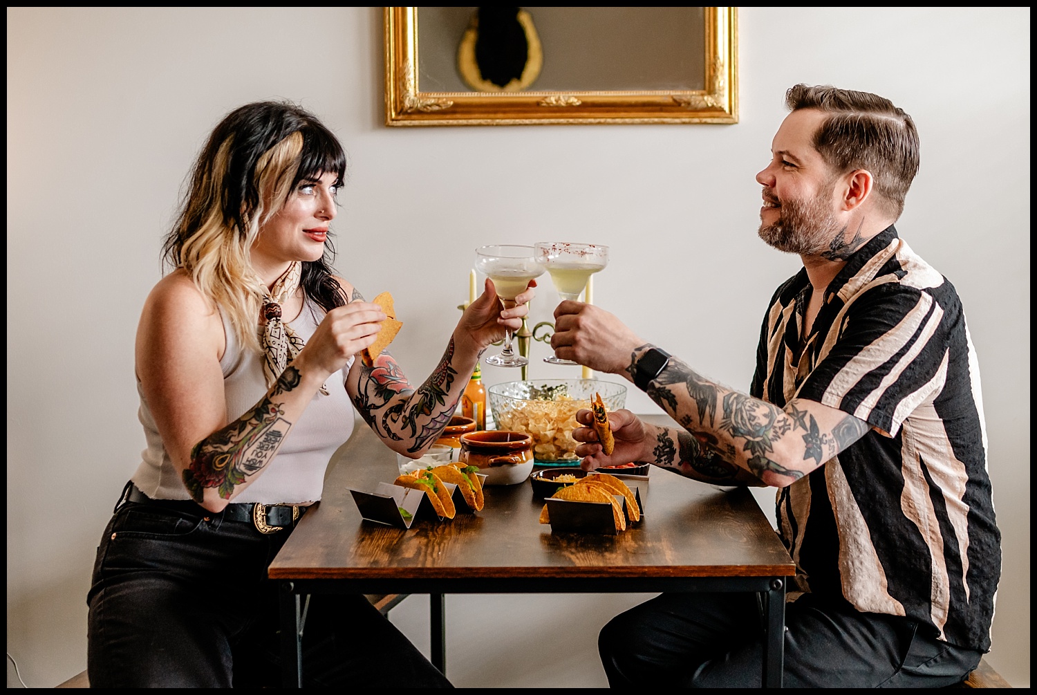 A woman and a man sit across from each other at a table, clinking margarita glasses during an in-home couples session. They smile over tacos, chips, and salsa; both have tattoos and casual clothing. A framed mirror hangs on the wall behind them.