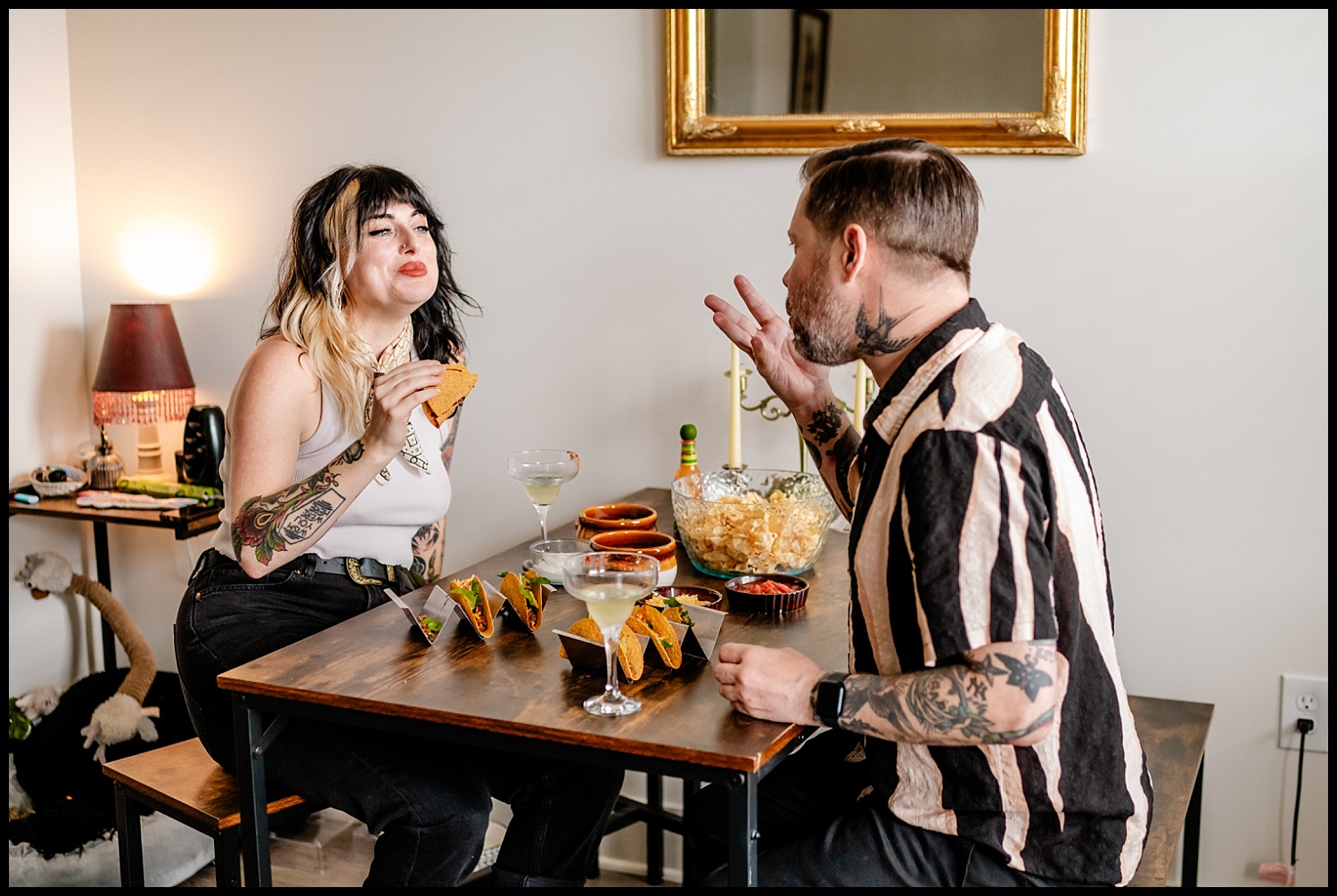 A tattooed couple enjoys tacos, drinks, and chips during a cozy Nashville couples session. They smile and laugh together in a warm room with soft lighting and a gold-framed mirror on the wall.