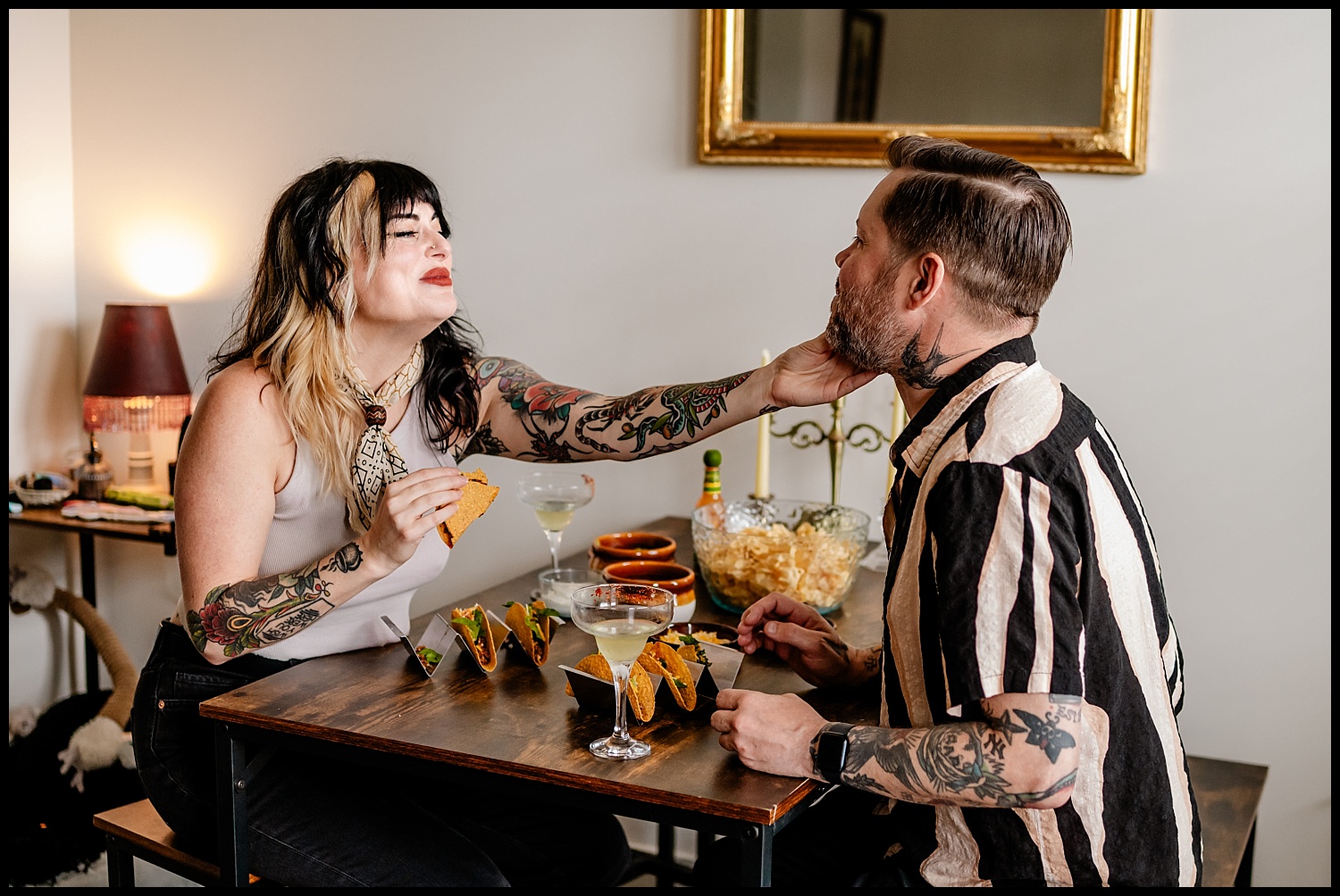A tattooed woman and man sit at a small table with tacos and drinks during a cozy Nashville couples session. She gently touches his face, smiling, while he leans forward. The scene includes snacks and charming decor in the background.