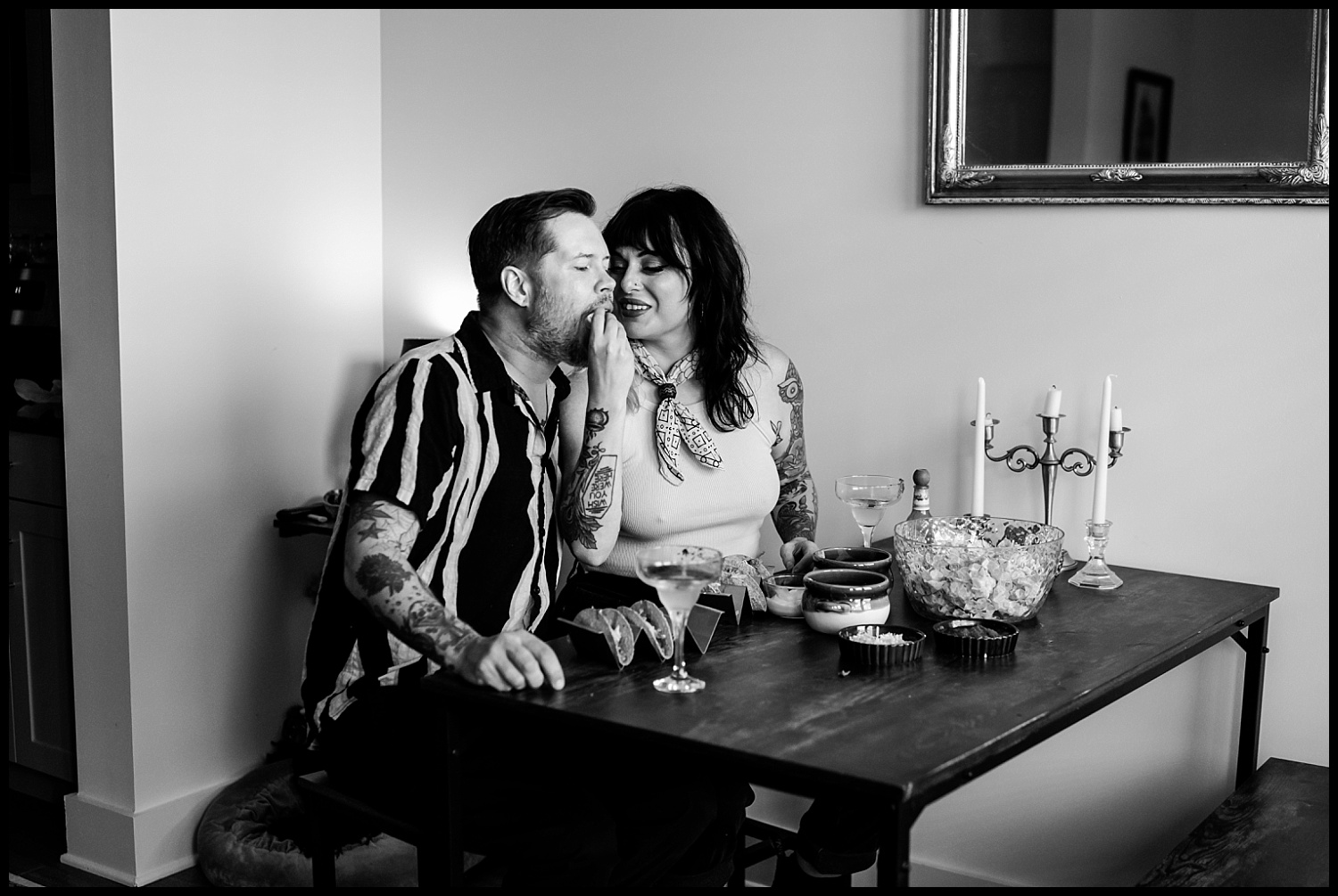 A tattooed couple sits closely at a dining table during an in-home couples session in Nashville, smiling as the woman whispers into the mans ear. Drinks, snacks, tacos, a candelabra, and a decorative bowl create an intimate and cheerful atmosphere.