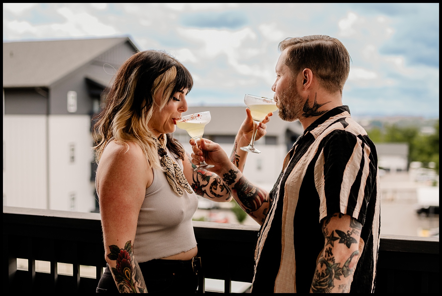 A tattooed man and woman stand on a balcony in Nashville, smiling and clinking cocktail glasses during their couples session. They are dressed casually, with modern buildings and a cloudy sky in the background—perfect for an in-home photography vibe.
