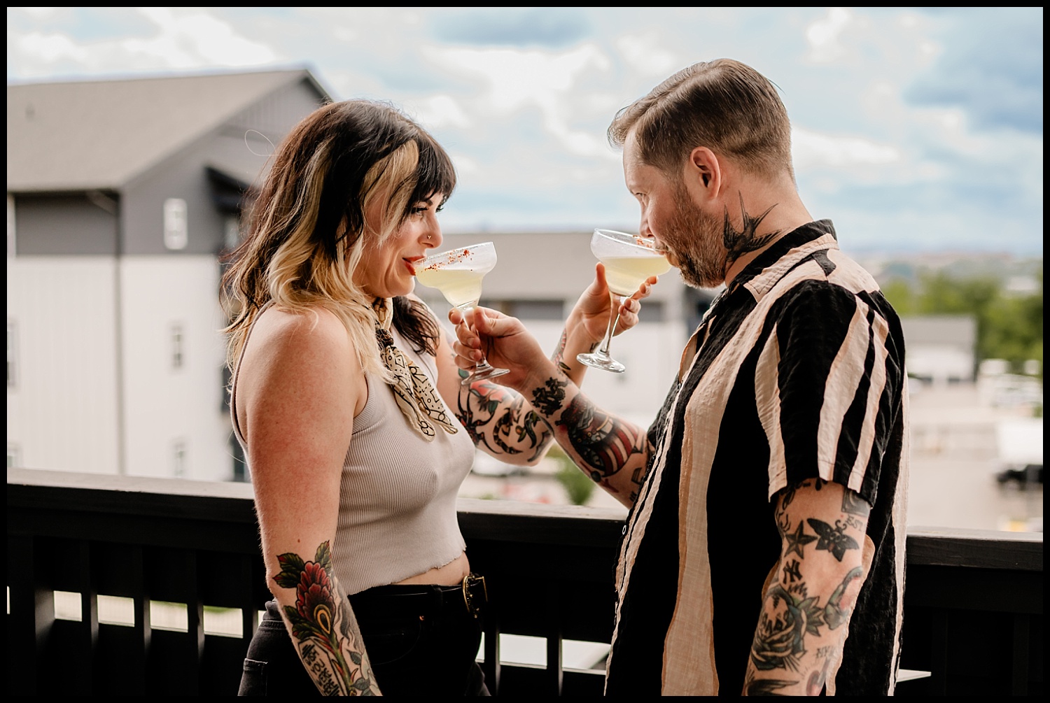 A woman and a man with tattoos clink cocktail glasses while standing close together on an outdoor balcony in Nashville, smiling at each other during their couples session, with buildings and a cloudy sky in the background.
