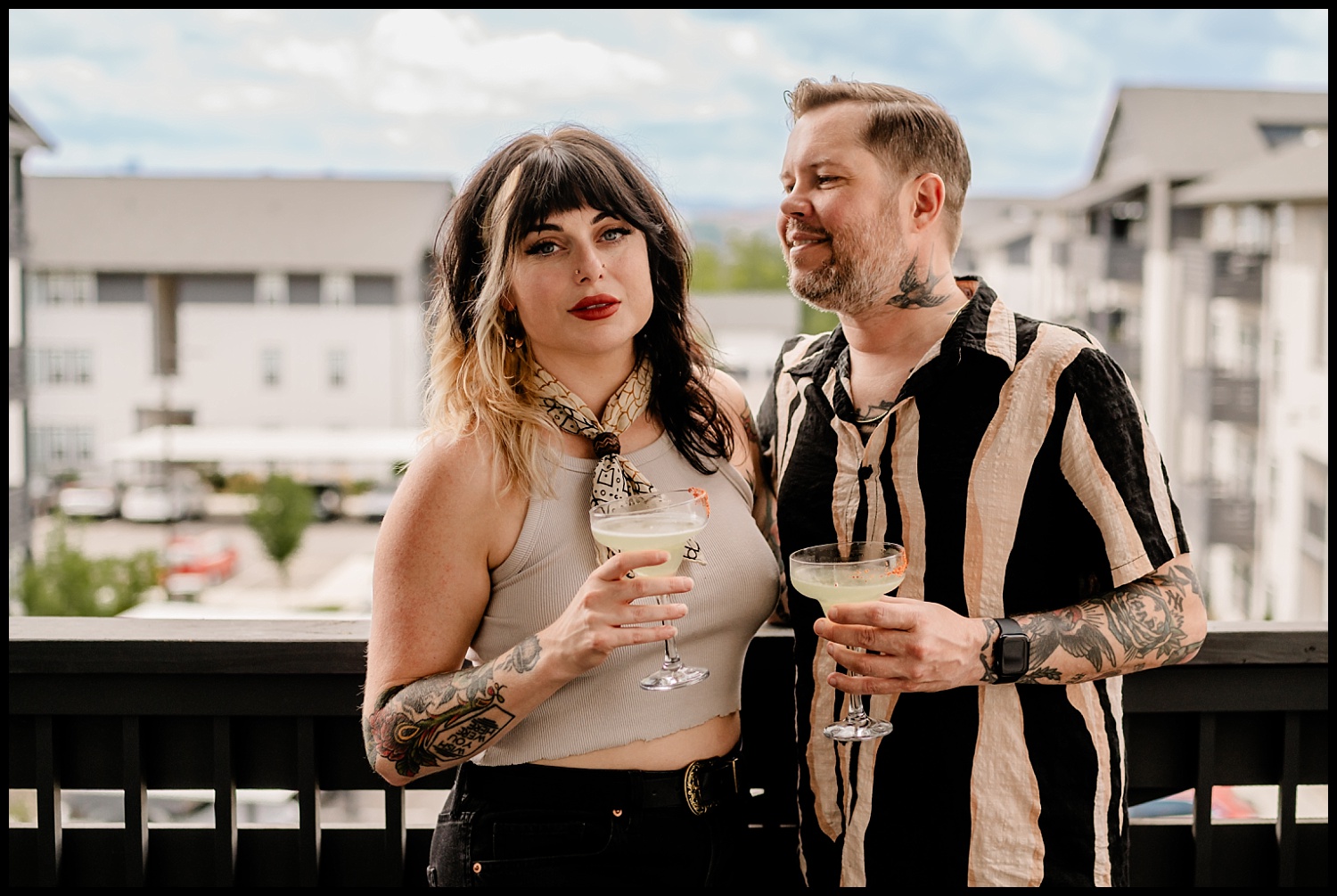A woman and a man with tattoos stand close together on a balcony during an in-home couples session in Nashville, holding cocktails and smiling. Apartment buildings and a cloudy sky are visible in the background.