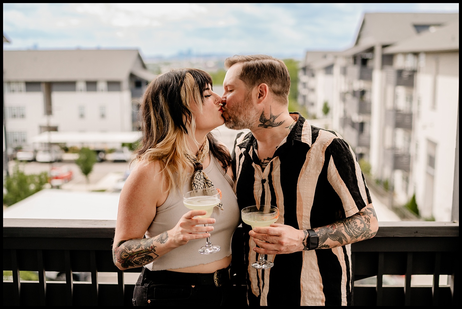 A couple stands on a balcony, kissing and holding cocktails—maybe tequila—in hand. Both have tattoos; the woman wears a light tank top and scarf, the man a striped shirt. Apartment buildings frame this intimate couples session.