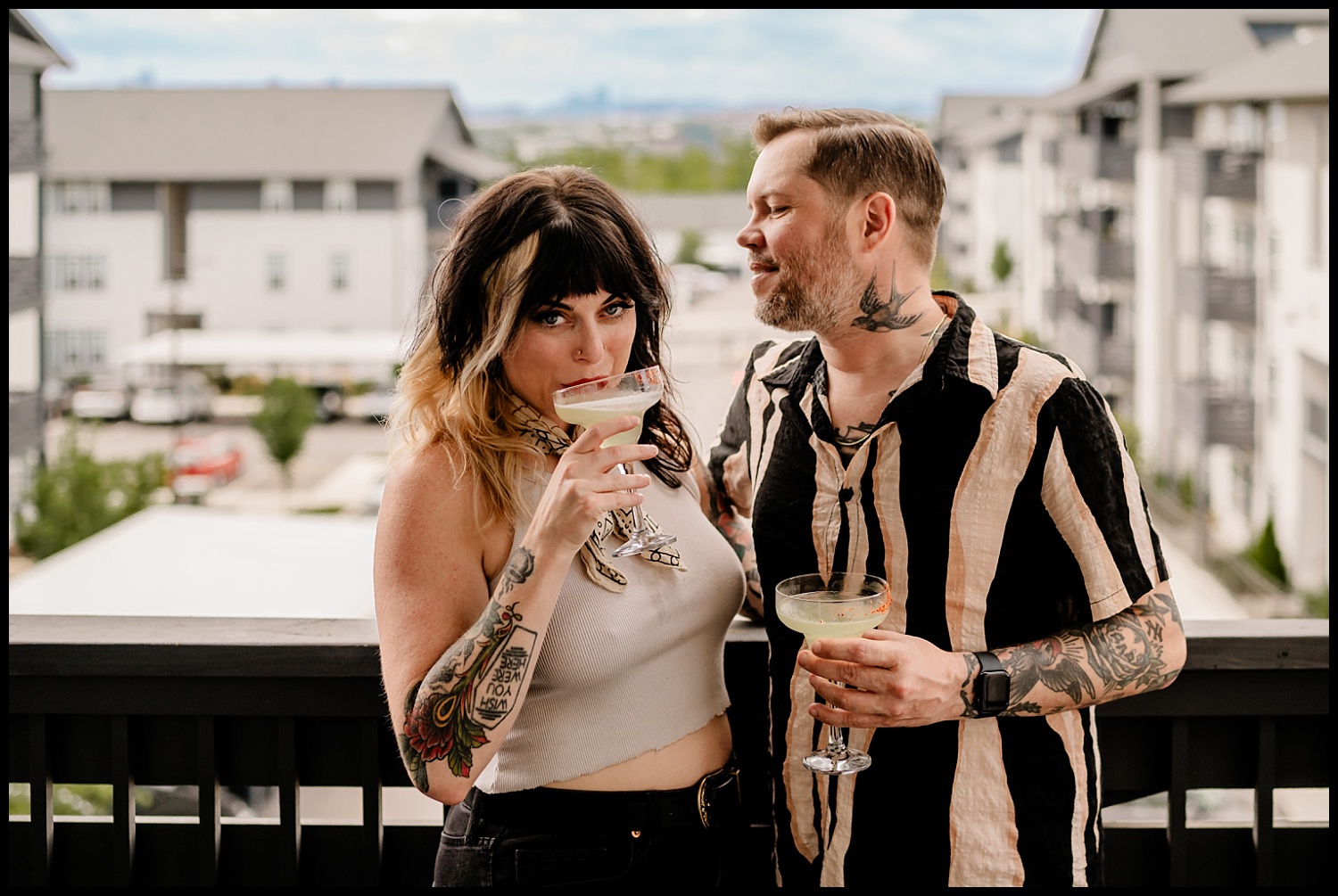 A woman with tattoos sips a cocktail while a man in a striped shirt stands beside her, both enjoying their drinks during a Nashville couples session on a balcony with modern apartment buildings in the background.