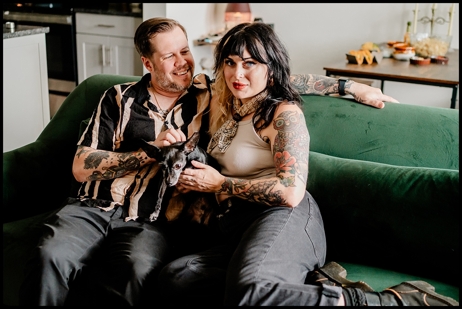 A tattooed couple sits close together on a green couch in their Nashville home, smiling and holding a small black dog. This cozy in-home photography session features a table with food and drinks visible in the background.