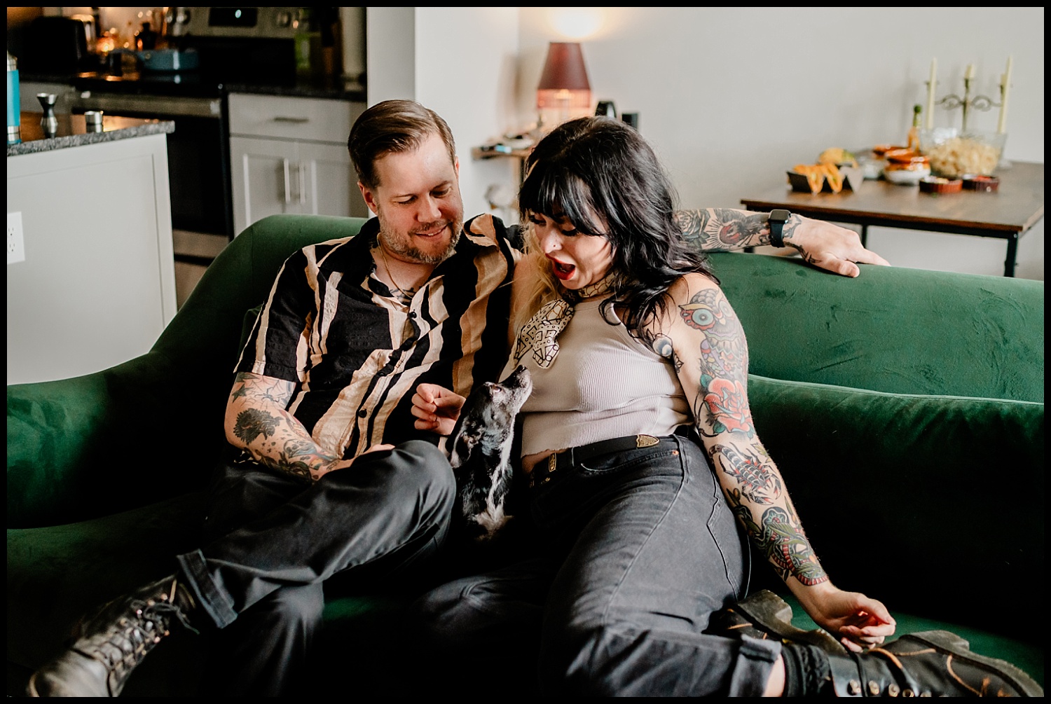 A man and woman with tattoos sit closely on a green couch, smiling and interacting with a small black and white dog on the womans lap during a cozy Nashville couples session in their inviting living room.