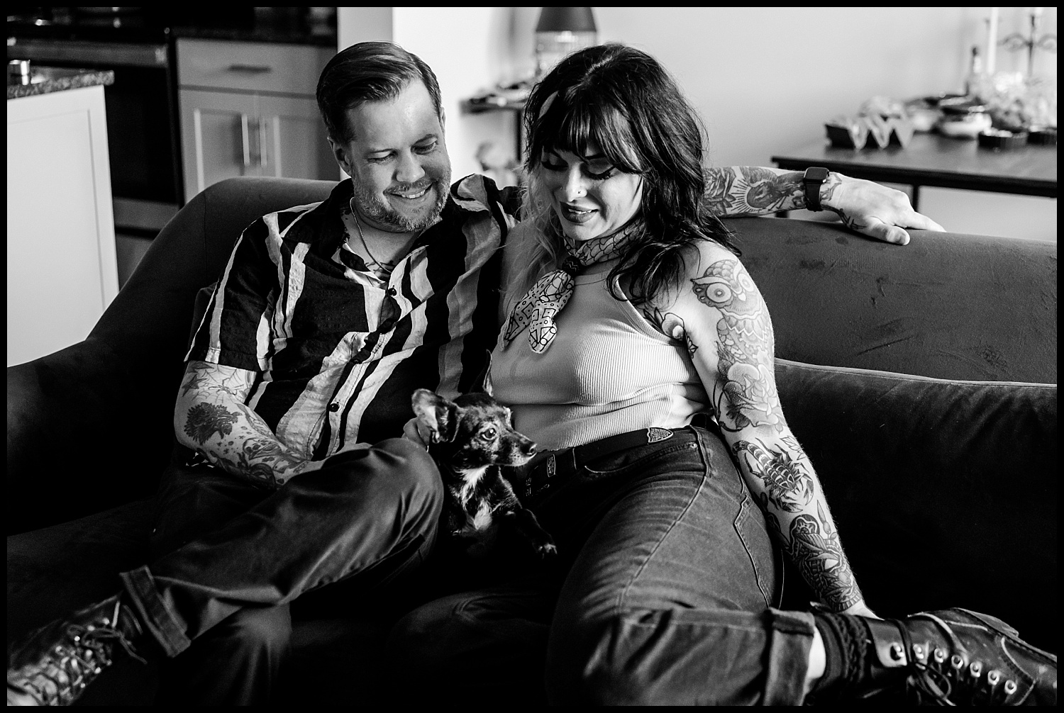 A tattooed couple sits closely together on a couch during an in-home couples session in Nashville, smiling at each other, with a small dog resting on the woman’s lap. The black and white image has a cozy, relaxed atmosphere.