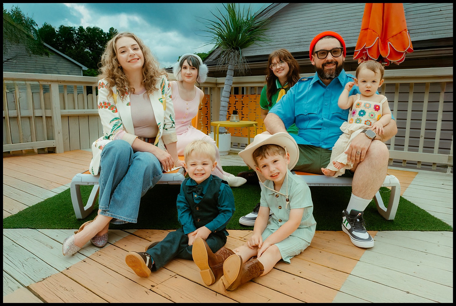 A smiling family of seven poses on a wooden deck for whimsical Family Portraits. Two adults and five children, in colorful, playful outfits, sit and kneel together amid greenery, a fence, and an orange umbrella—evoking subtle Wes Anderson vibes.
