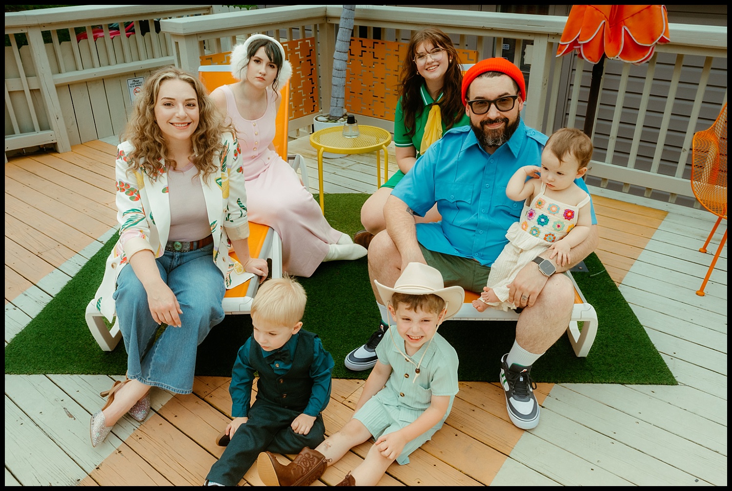 A group of six adults and two young children pose together on a deck in Nashville. Some sit or crouch on artificial grass; everyone is smiling and casually dressed in bright, retro-style clothing, evoking the charm of Wes Anderson family portraits.