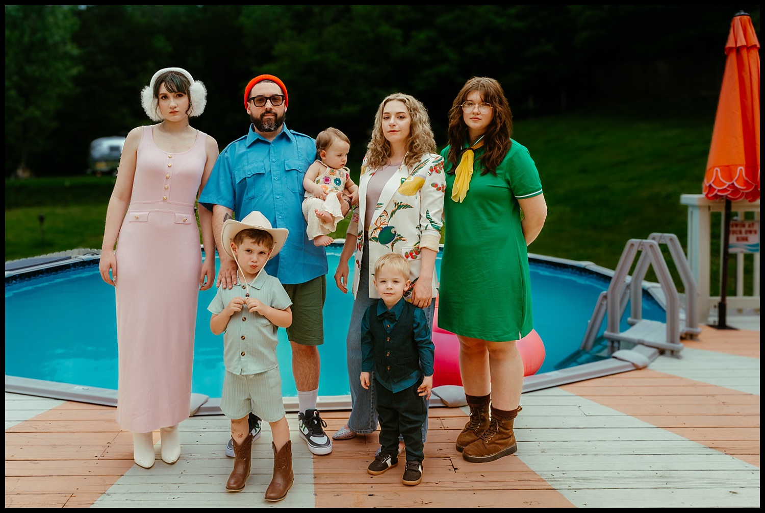 Six people and two children stand on a deck by a backyard pool, channeling Wes Anderson vibes with colorful, retro-inspired clothing. Perfect for Family Portraits, the group gazes at the camera amid trees and grass—Nashville charm in the background.