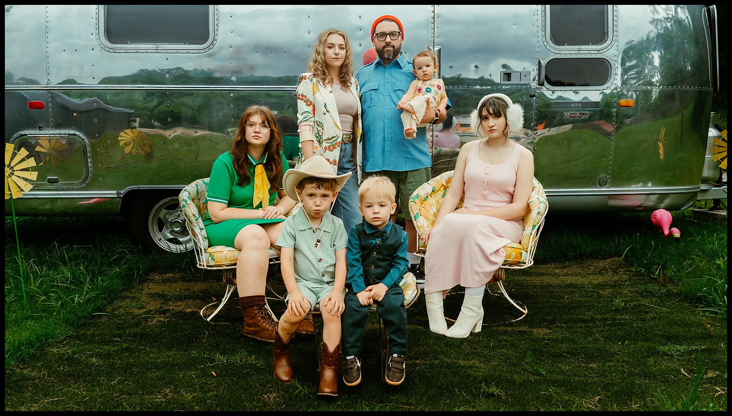 A family of seven poses Wes Anderson–style in front of a shiny silver camper. The adults stand behind; four children sit or stand in front, all in colorful, retro-inspired outfits and boots, with green grass and yellow chairs nearby.