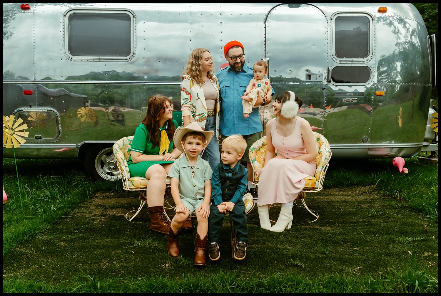 A family of seven poses outdoors in front of a shiny silver Airstream, dressed in colorful, vintage-style clothing. Their arrangement and style bring to mind Wes Anderson family portraits with a Nashville flair.