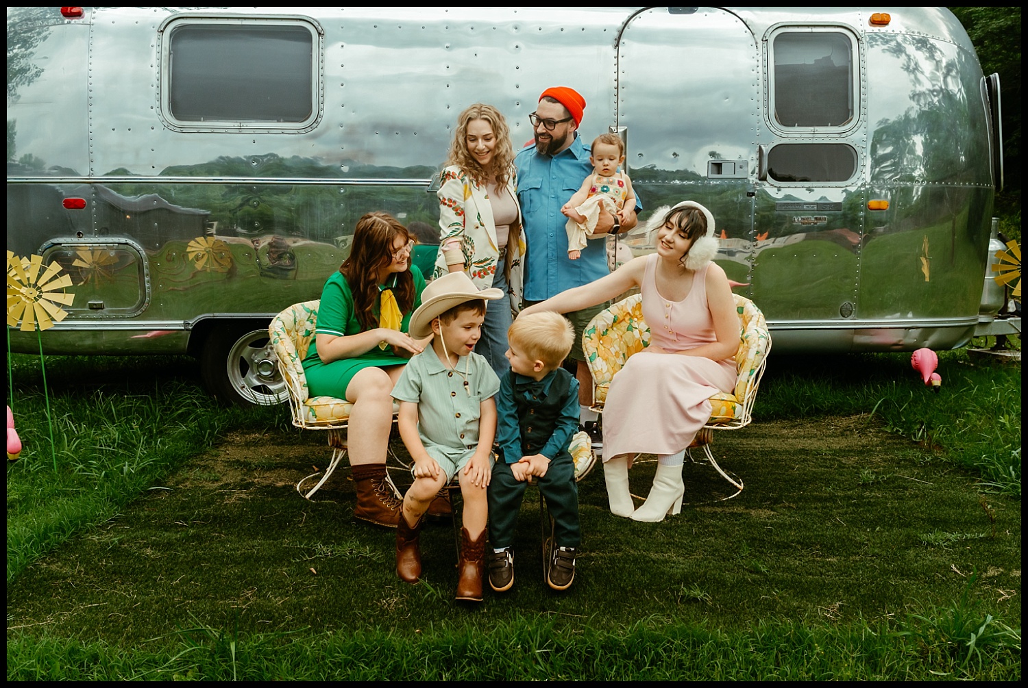 A group of adults and children sit and stand on vintage chairs in front of a shiny silver Airstream trailer outdoors, smiling and interacting warmly in a scene reminiscent of Wes Anderson family portraits near Nashville.