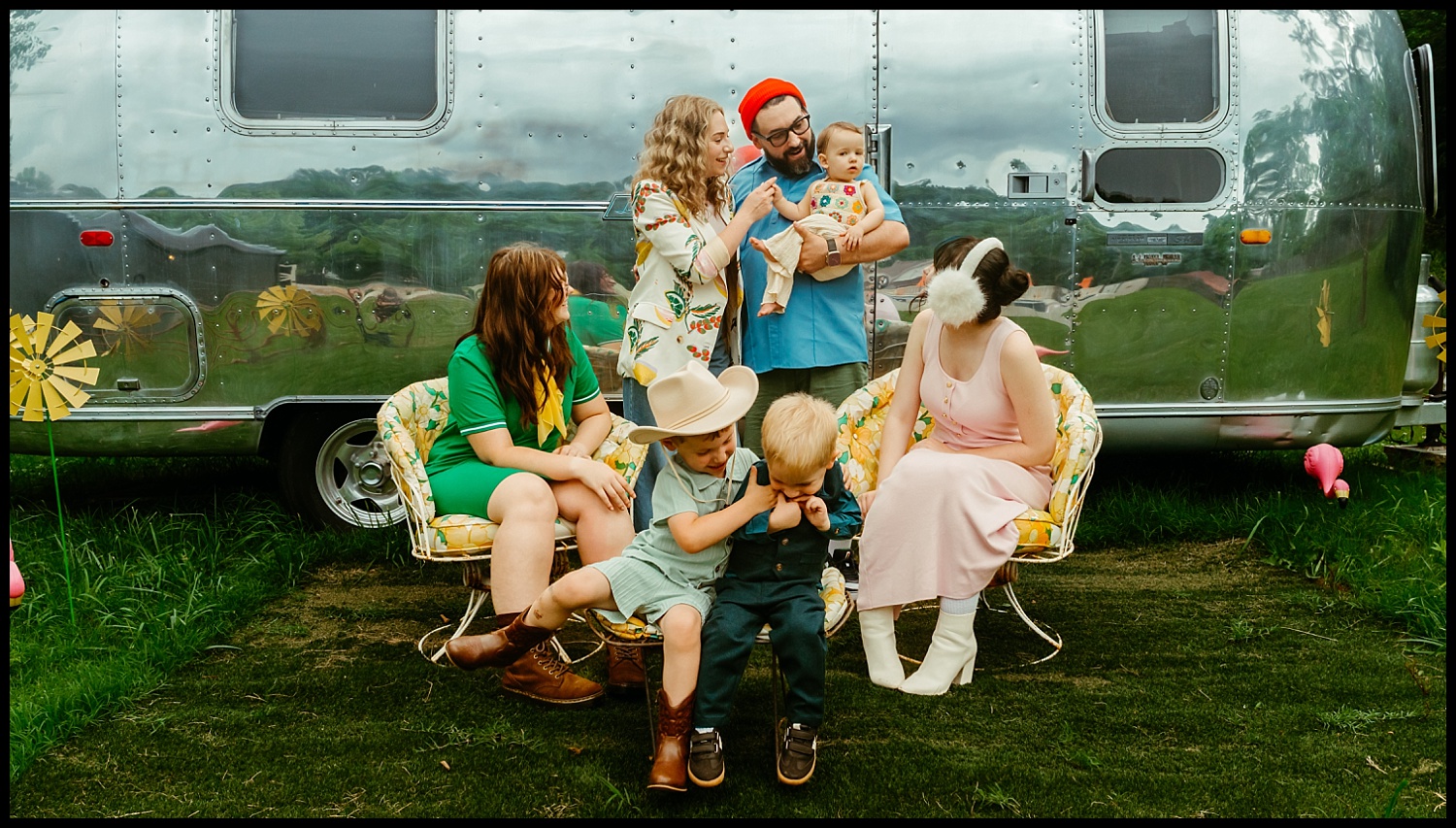 Six people, posed like a Wes Anderson family portrait, sit and stand in front of a shiny silver trailer in Nashville. The adults smile warmly as the children, seated on a floral bench on grass, share playful expressions.