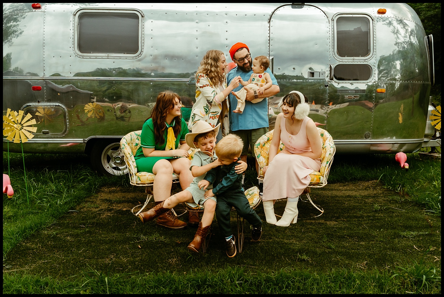 A group of six adults and two children sit and stand in front of a shiny silver trailer on grass, smiling and interacting with each other. With colorful clothing and chairs, the scene channels Wes Anderson vibes—perfect for Nashville family portraits.
