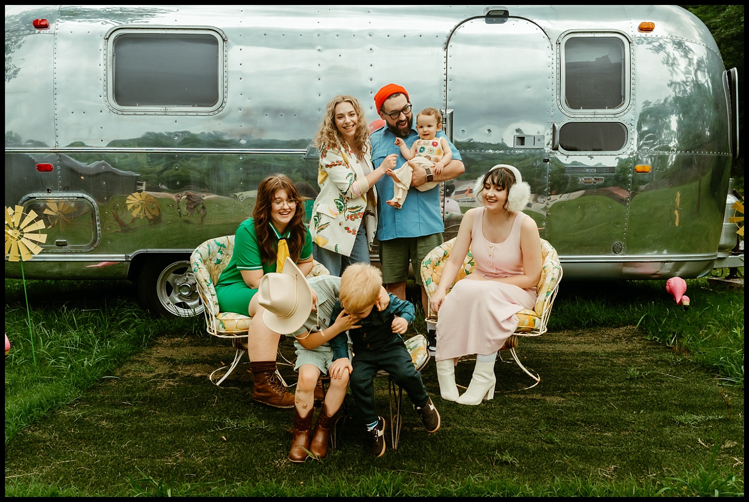 Six people, including two children, pose and smile in front of a shiny silver Airstream trailer. Dressed in retro clothing, they create a cheerful scene that feels straight out of a Wes Anderson film—quirky vibes for unforgettable family portraits outdoors.